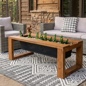May include: A rectangular wooden coffee table featuring a built-in black metal trough filled with green glass bottles. The table has a natural wood finish with a thick frame and open sides, positioned on a patterned gray and white area rug.