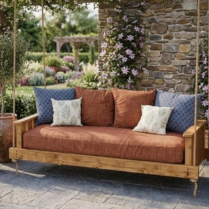 May include: A wooden porch swing featuring a long, rust-colored cushion and a collection of throw pillows. The pillows include two large blue patterned squares, two large rust-colored squares, and two smaller white squares with green leaf prints.