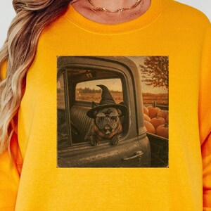 May include: A bright yellow crewneck sweatshirt featuring a central graphic print of a dog wearing a witch hat and glasses, sitting in the window of a vintage truck parked near a pumpkin patch during a sunset, with long sleeves and ribbed cuffs.