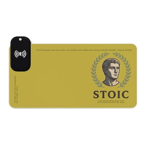 May include: A rectangular mustard yellow desk mat featuring a black wireless charging pad on the left. The mat displays a portrait of a man in a laurel wreath, the word STOIC, and quotes from Marcus Aurelius and Seneca in black text.