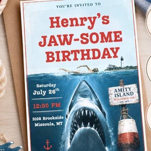 May include: A rectangular birthday invitation card featuring a shark emerging from the water, a swimmer, a lighthouse, and a sign reading AMITY ISLAND WELCOMES YOU EST. 1975. The text reads YOU'RE INVITED TO Henry's JAW-SOME BIRTHDAY, Saturday July 26th, 12:00 PM, 3108 Brookside Missoula, MT.