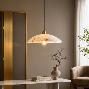 Może przedstawiać: Lampa wisząca z szerokim, płytkim kloszem w kształcie kopuły z białego marmuru z szarymi żyłkami, zawieszona na czarnym przewodzie przy mosiężnym mocowaniu. Lampa zawiera pojedynczą żarówkę żarnikową umieszczoną pod marmurową kopułą.
