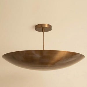 May include: A ceiling-mounted light fixture featuring a wide, shallow, bowl-shaped shade with a brushed bronze finish. The shade is suspended by a single vertical rod connected to a circular canopy that attaches to the ceiling surface.