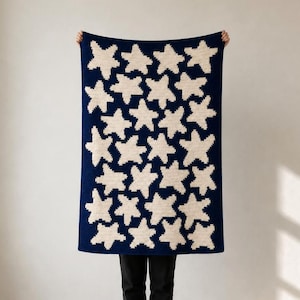 Könnte beinhalten: Eine rechteckige Strickdecke mit einem sich wiederholenden Muster aus cremefarbenen Sternen auf einem tief dunkelblauen Hintergrund. Das Textil zeigt eine konsistente, gitterartige Anordnung der Sternmotive über die gesamte Stoffoberfläche.