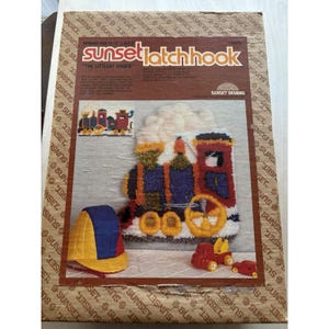 May include: A Sunset Latch Hook kit box titled The Littlest Engine, featuring a finished latch hook train design with white smoke, a blue, red, and yellow engine, a matching yellow and blue cap, and two small red toy trucks on the packaging.