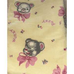 May include: A pale yellow blanket featuring a repeating pattern of grey bear heads with pink bows. The bears are winking and smiling. Small pink and blue flowers, butterflies, and the word "Wuzzy" in pink script are scattered across the fabric.