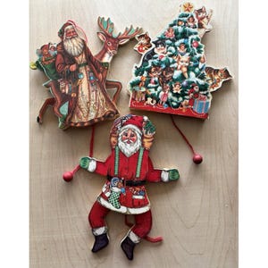 May include: Three vintage-style Christmas ornaments featuring a Santa Claus with a reindeer, a decorated Christmas tree with kittens, and a jointed Santa Claus figure with red clothing, green suspenders, and red wooden beads on strings.