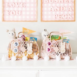 Può includere: Due cesti in vimini rosa pastello pieni di regali e giocattoli. I cesti sono decorati con orecchie di coniglio e cartellini con nomi personalizzati. I cesti sono su una superficie bianca con una fila di pompon bianchi.
