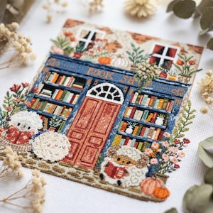 Può includere: Un ricamo a punto croce che raffigura una vetrina blu con la scritta BOOK SHOP sopra una porta centrale arancione. Il disegno presenta scaffali pieni di libri colorati, due piccole pecore che leggono, zucche e accenti floreali.