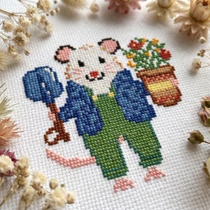 May include: A cross-stitch embroidery on white fabric featuring a white mouse wearing a blue jacket and green overalls. The mouse holds a blue shovel in one hand and a brown flower pot with red and yellow flowers in the other hand.