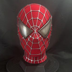 May include: A red full-head mask featuring a raised silver web pattern across the entire surface. The mask includes two large, angular, metallic silver eye lenses with a smooth finish, designed to cover the eyes completely in a classic superhero style.