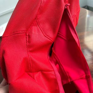 May include: A bright red textured fabric item featuring a visible zipper closure along the side. The material has a distinct grid-like pattern across the surface, with smooth red fabric visible on the interior side of the open zipper seam.