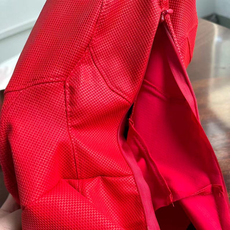 May include: A bright red textured fabric item featuring a visible zipper closure along the side. The material has a distinct grid-like pattern across the surface, with smooth red fabric visible on the interior side of the open zipper seam.