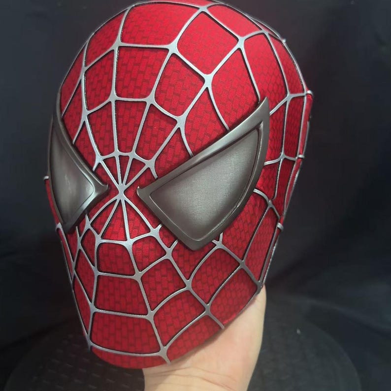 May include: A red full-head mask featuring a textured surface with a prominent silver web pattern overlay. The mask includes two large, angular, metallic-toned eye lenses with a smooth finish, covering the entire face area in a classic superhero style.