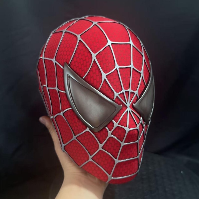 May include: A red full-head mask featuring a raised silver web pattern across the surface and two large, angular, metallic silver eye lenses. The fabric has a fine, textured honeycomb-like detail throughout the red sections of the mask.