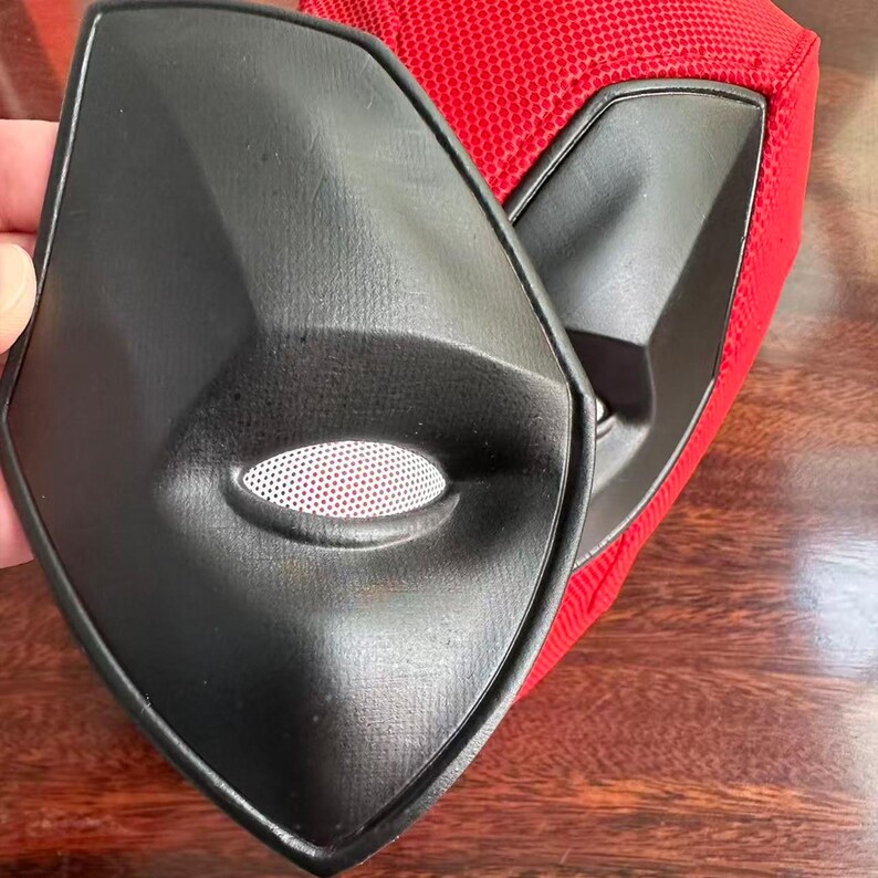 May include: A black, angular eye mask component with a white mesh eye opening, positioned in front of a red, textured fabric mask base. The black piece features a smooth, matte finish and a sharp, geometric shape designed for facial coverage.