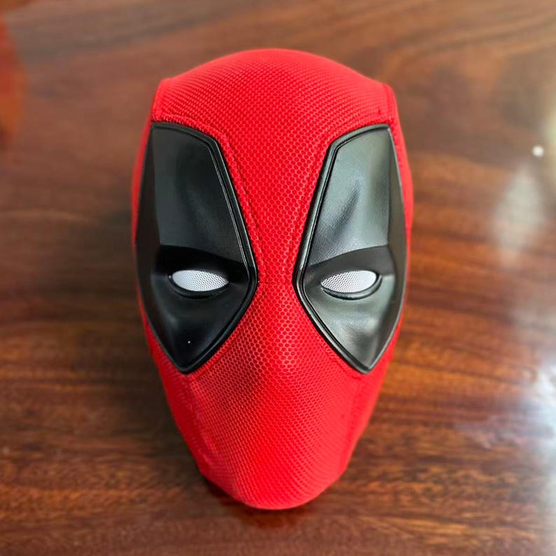 May include: A red fabric mask featuring a textured surface and two large, black, angular eye patches with white mesh lenses. The mask is designed to cover the head and face, showcasing a distinct, molded shape with prominent black eye contours.
