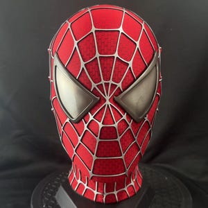 May include: A red full-head mask featuring a raised silver web pattern across the entire surface. The mask includes two large, angular, metallic silver eye lenses with a smooth, reflective finish, covering the eye area in a distinct shape.