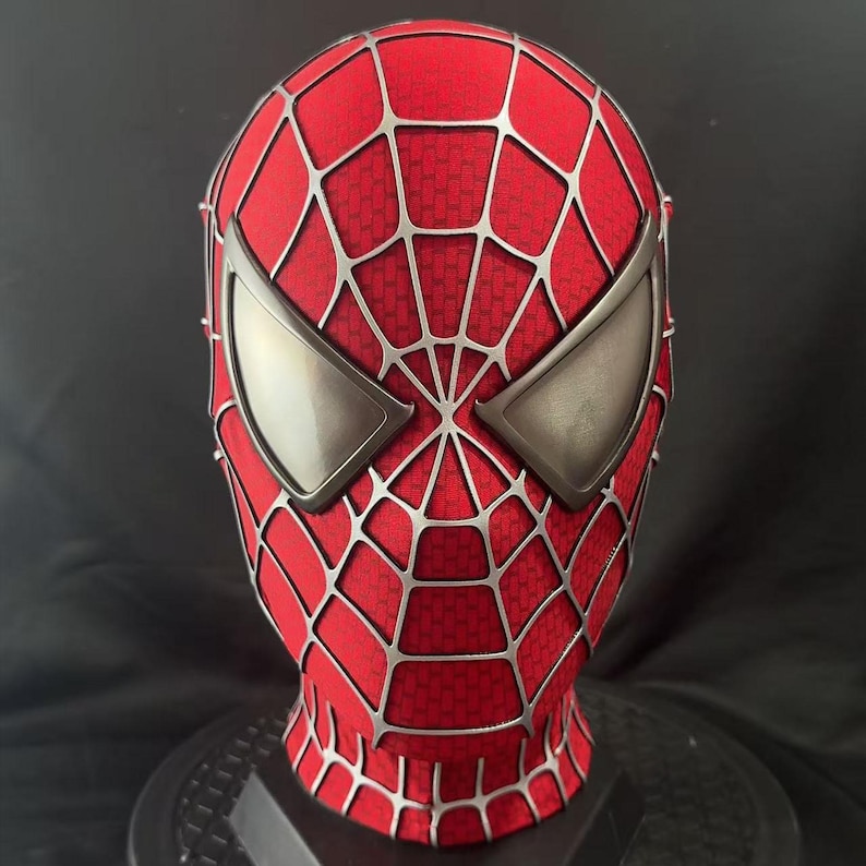 May include: A red full-head mask featuring a raised silver web pattern across the entire surface. The mask includes two large, angular, metallic silver eye lenses with a smooth, reflective finish, covering the eye area in a distinct shape.