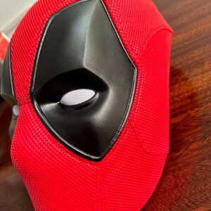 May include: A red full-face mask featuring a textured, honeycomb-like surface pattern. The mask includes a prominent, smooth black eye section with a white mesh insert over the eye opening, creating a distinct contrast against the vibrant red material.