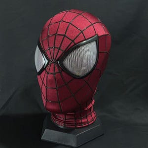 Pode incluir: Uma máscara de cabeça inteira com uma base de tecido vermelho escuro e um padrão de teia de aranha preto. A máscara inclui duas lentes oculares grandes e angulares com textura de malha branca, emolduradas por bordas pretas, num suporte.
