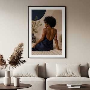 May include: A framed wall art print featuring a person with dark curly hair seen from behind, wearing a dark blue satin slip dress with thin straps. The person is seated on a bed next to a blue and white patterned vase with green foliage.