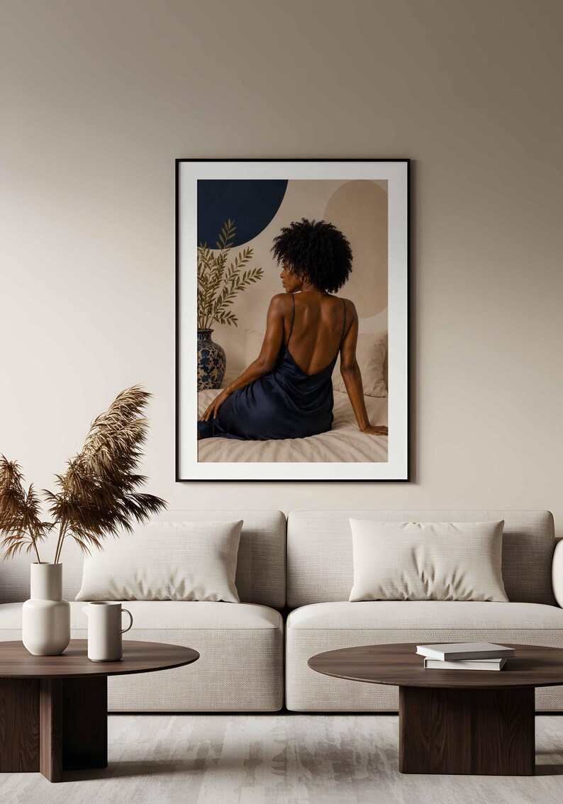 May include: A framed wall art print featuring a person with dark curly hair seen from behind, wearing a dark blue satin slip dress with thin straps. The person is seated on a bed next to a blue and white patterned vase with green foliage.