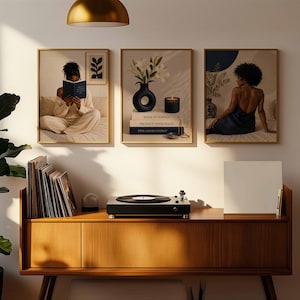 May include: Three framed wall art prints displayed above a wooden media console. The left print shows a person reading a book, the center print features a vase with white flowers and stacked books, and the right print depicts a person sitting.