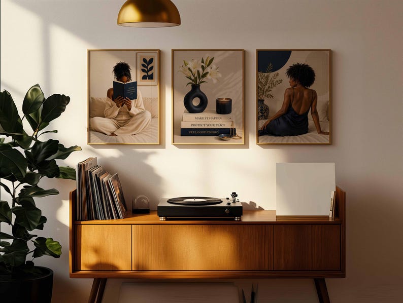May include: Three framed wall art prints displayed above a wooden media console. The left print shows a person reading a book, the center print features a vase with white flowers and stacked books, and the right print depicts a person sitting.