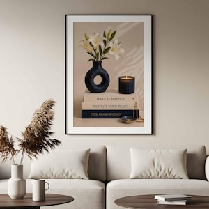 May include: A framed wall art print featuring a black donut-shaped vase with white lily flowers, a dark ribbed candle, and a stack of three books labeled MAKE IT HAPPEN, PROTECT YOUR PEACE, and FEEL GOOD ENERGY, with a small bowl and gold chain.