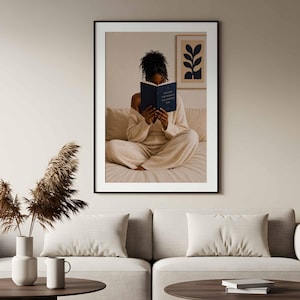 May include: A framed wall art print featuring a person with dark curly hair sitting cross-legged on a bed, reading a dark blue book with the text "BECOME THE WOMAN YOU PRAYED FOR" on the cover. The print includes a small framed botanical art piece.