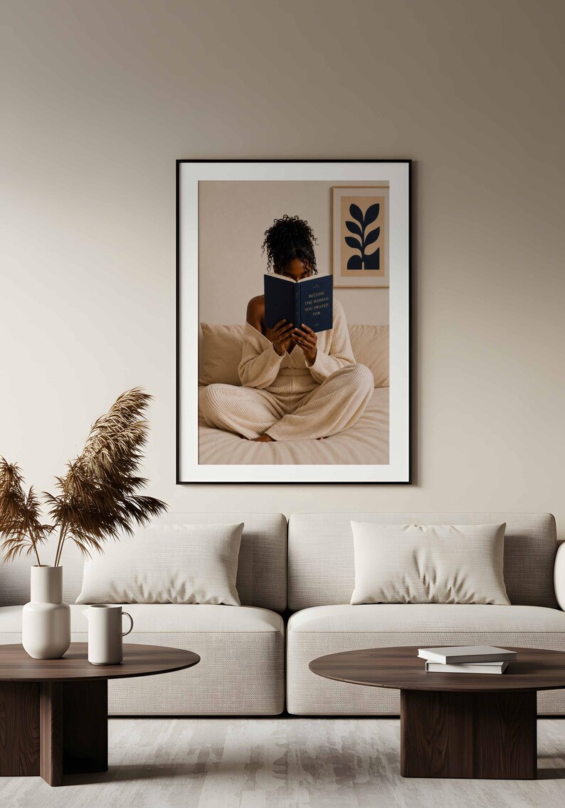 May include: A framed wall art print featuring a person with dark curly hair sitting cross-legged on a bed, reading a dark blue book with the text "BECOME THE WOMAN YOU PRAYED FOR" on the cover. The print includes a small framed botanical art piece.