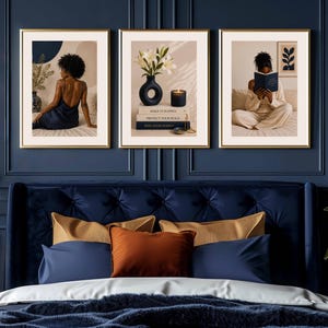 May include: A set of three framed wall art prints featuring a person in a dark dress, a vase of white lilies with books and a candle, and a person reading a book. The prints are displayed in gold frames against a dark blue paneled wall.