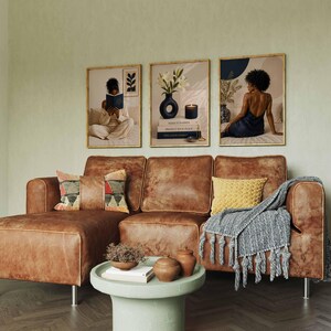 May include: A brown leather sectional sofa with a patterned pillow, a yellow textured pillow, and a gray tasseled throw blanket. A round light green coffee table sits in front with two small bowls and books. Three framed wall art prints.