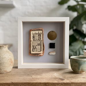 May include: A square white shadow box frame containing a collage of a vintage black and white illustration of a cabinet on a textured brown backing, a gold circular disc, a small black square, and a thin white rectangular piece on a grey background.