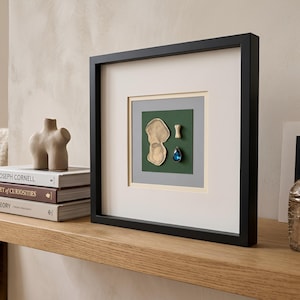 May include: A square black frame containing a green mount with three mounted objects: a textured gold-toned organic shape, a small white bone-like piece, and a teardrop-shaped pendant with a deep blue stone set in a silver-coloured metal frame.