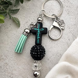 May include: A keychain featuring a black cross pendant with the word Jesus written in teal script. The design includes a light teal suede tassel, a large black textured bead, a clear faceted bead, and a silver metal bead on a silver ring.