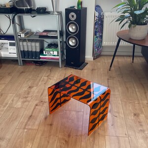 Könnte beinhalten: Ein rechteckiger Beistelltisch aus durchscheinendem orangefarbenem Acrylmaterial mit einem sich wiederholenden schwarzen Tigerstreifenmuster auf der gesamten Oberfläche. Der Tisch hat ein Wasserfall-Design mit glatten, abgerundeten Kanten.