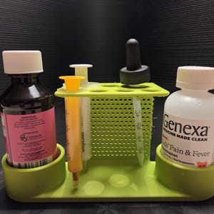 May include: A lime green plastic organizer stand with two circular bottle holders and a central perforated rack. It holds a dark bottle with a white cap, a yellow oral syringe, a clear oral syringe, a dropper, and a Genexa Kids' Pain & Fever bottle.