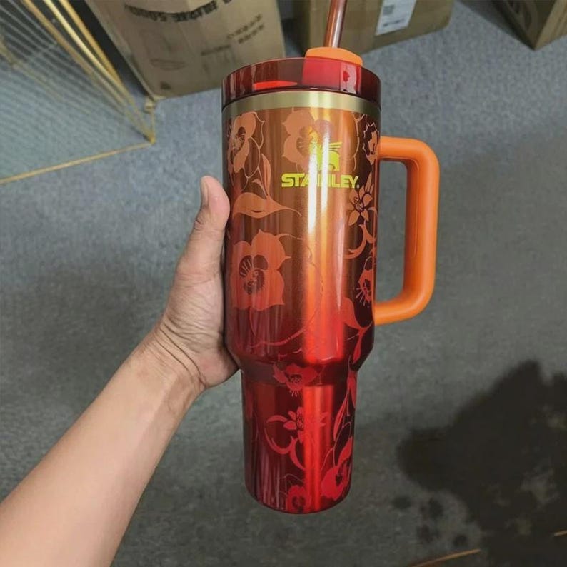 May include: A metallic orange Stanley tumbler with a floral pattern, featuring a large orange side handle, a matching orange lid, and a clear straw. The brand name Stanley is printed in yellow on the upper front of the cylindrical vessel.