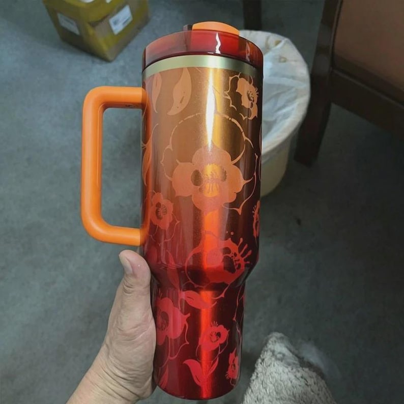 May include: A tall, cylindrical metal tumbler with a gradient finish transitioning from deep red at the base to a lighter orange at the top. It features a repeating floral pattern, a bright orange side handle, and a matching orange lid.
