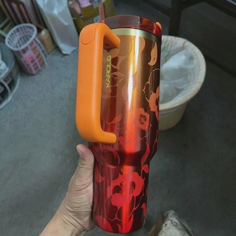 May include: A tall, cylindrical stainless steel tumbler with a gradient orange and red finish featuring a floral pattern. It has a large, solid orange handle attached to the side, a dark lid, and the word KAROLG printed vertically in yellow.