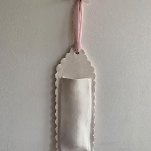 Op de afbeelding: Een witte keramische wandzak met een geschulpte rand en een gladde, glanzende afwerking. Het verticale, rechthoekige stuk heeft een afgeronde bovenkant met een gat om op te hangen. Een lichtroze stoffen lint is door het gat gestrikt.
