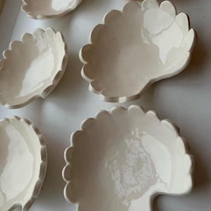 Könnte beinhalten: Eine Sammlung von cremeweißen Keramikschalen in Form von gewellten Muscheln. Jede Schale hat eine glatte, glänzende Oberfläche mit einem markanten geschwungenen Rand und einer flachen, abgerundeten Vertiefung, die in einer Gruppe angeordnet sind.