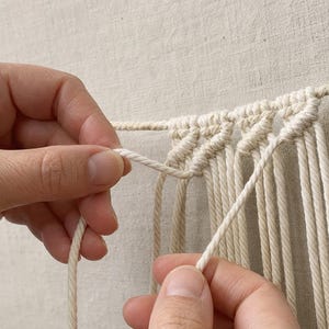 Pu&ograve; includere: Corda in cotone bianco sporco disposta in una serie di nodi a festone diagonali. La corda &egrave; sospesa orizzontalmente e fissata con uno spillo, con diversi fili verticali che pendono come parte di un progetto di macram&egrave; in fase di lavorazione.