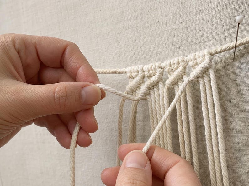 Pu&ograve; includere: Corda in cotone bianco sporco disposta in una serie di nodi a festone diagonali. La corda &egrave; sospesa orizzontalmente e fissata con uno spillo, con diversi fili verticali che pendono come parte di un progetto di macram&egrave; in fase di lavorazione.