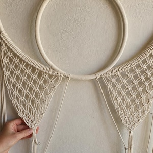 Pu&ograve; includere: Un arazzo da parete in macram&egrave; color crema caratterizzato da un grande cerchio avvolto in corda. Due sezioni triangolari di rete annodata complessa si estendono verso il basso dal cerchio, con lunghe frange verticali sciolte che pendono.
