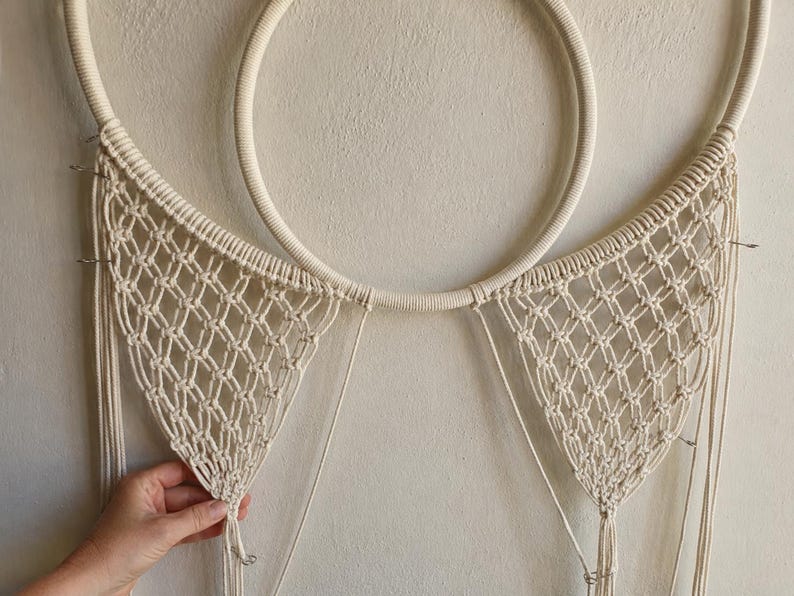 Pu&ograve; includere: Un arazzo da parete in macram&egrave; color crema caratterizzato da un grande cerchio avvolto in corda. Due sezioni triangolari di rete annodata complessa si estendono verso il basso dal cerchio, con lunghe frange verticali sciolte che pendono.