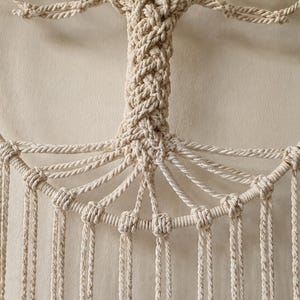 Pu&ograve; includere: Una decorazione da parete in macram&egrave; color crema con un motivo ad albero della vita. Il tronco centrale &egrave; realizzato in corda intrecciata e si estende in rami collegati a una cornice circolare. Numerose corde verticali pendono dalla base.