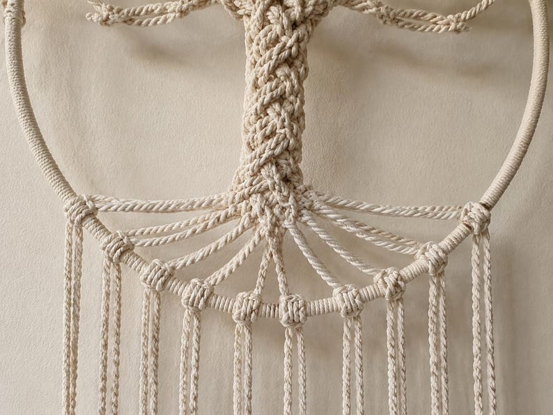 Pu&ograve; includere: Una decorazione da parete in macram&egrave; color crema con un motivo ad albero della vita. Il tronco centrale &egrave; realizzato in corda intrecciata e si estende in rami collegati a una cornice circolare. Numerose corde verticali pendono dalla base.